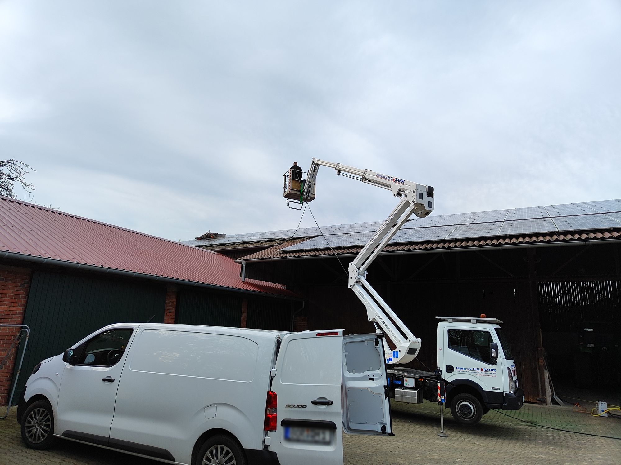 Hubarbeitsbühne mit einem Mitarbeiter im Korb an einem großen Hallendach mit Photovoltaikanlage, davor ein weißer Transporter.