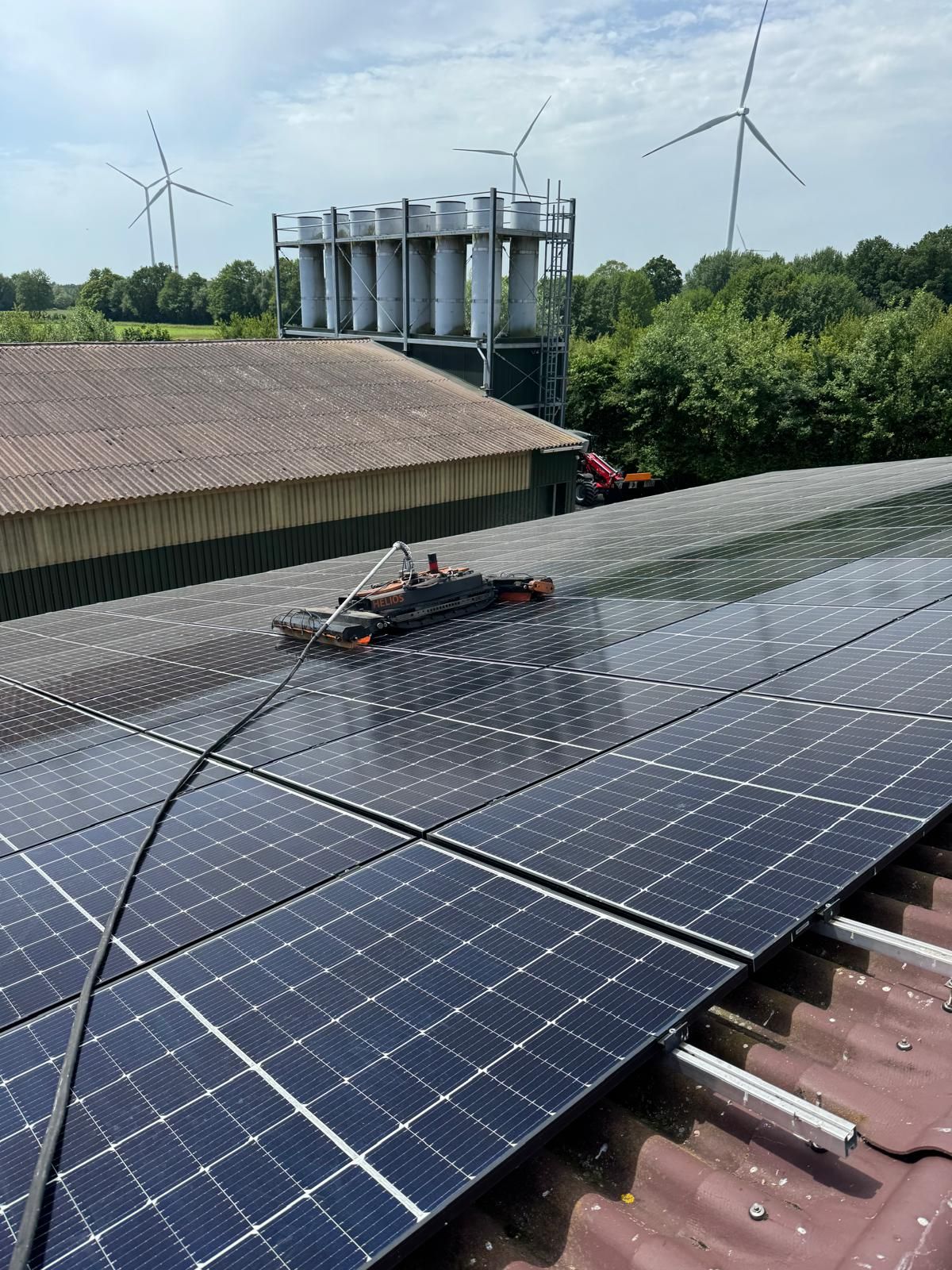 Reinigungsroboter auf einem großen PV-Stalldach mit nassen Modulen, dahinter eine Abluftanlage, Bäume und Windräder.